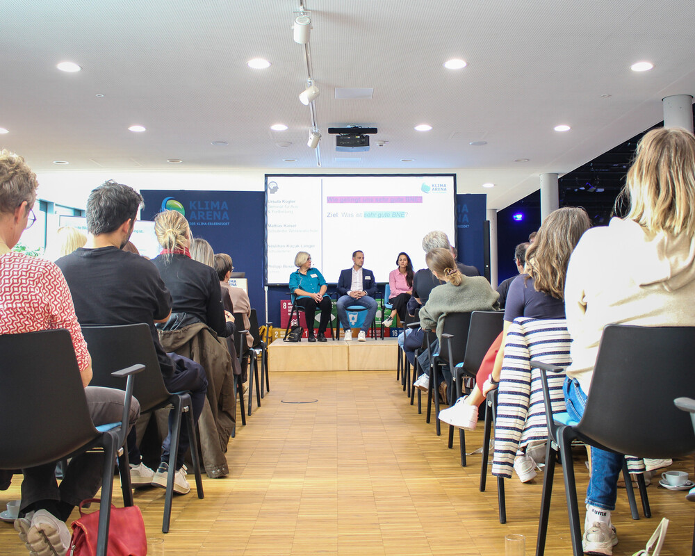 Blick durch die Stuhlreihen auf die Gäste einer Podiumsdiskussion auf der Bühne